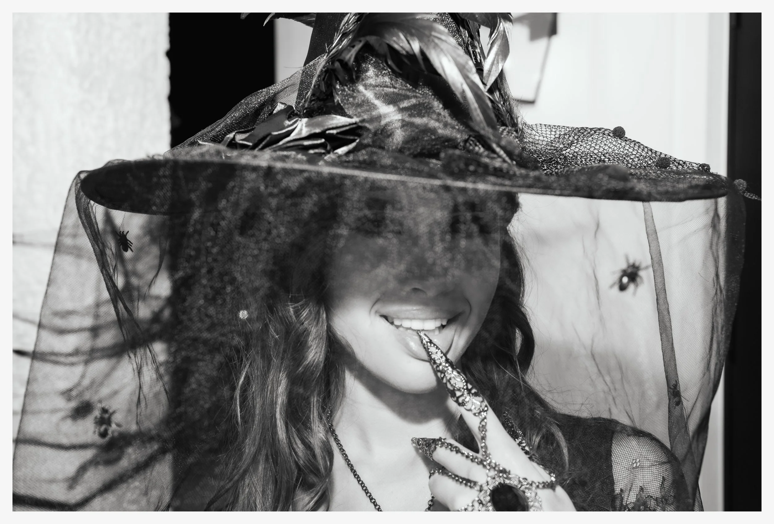 Woman dressed as a witch wearing a large, pointed hat and a lace veil with spider and insect motifs, smiling with her tongue out touching a decorative spider ring on her finger.