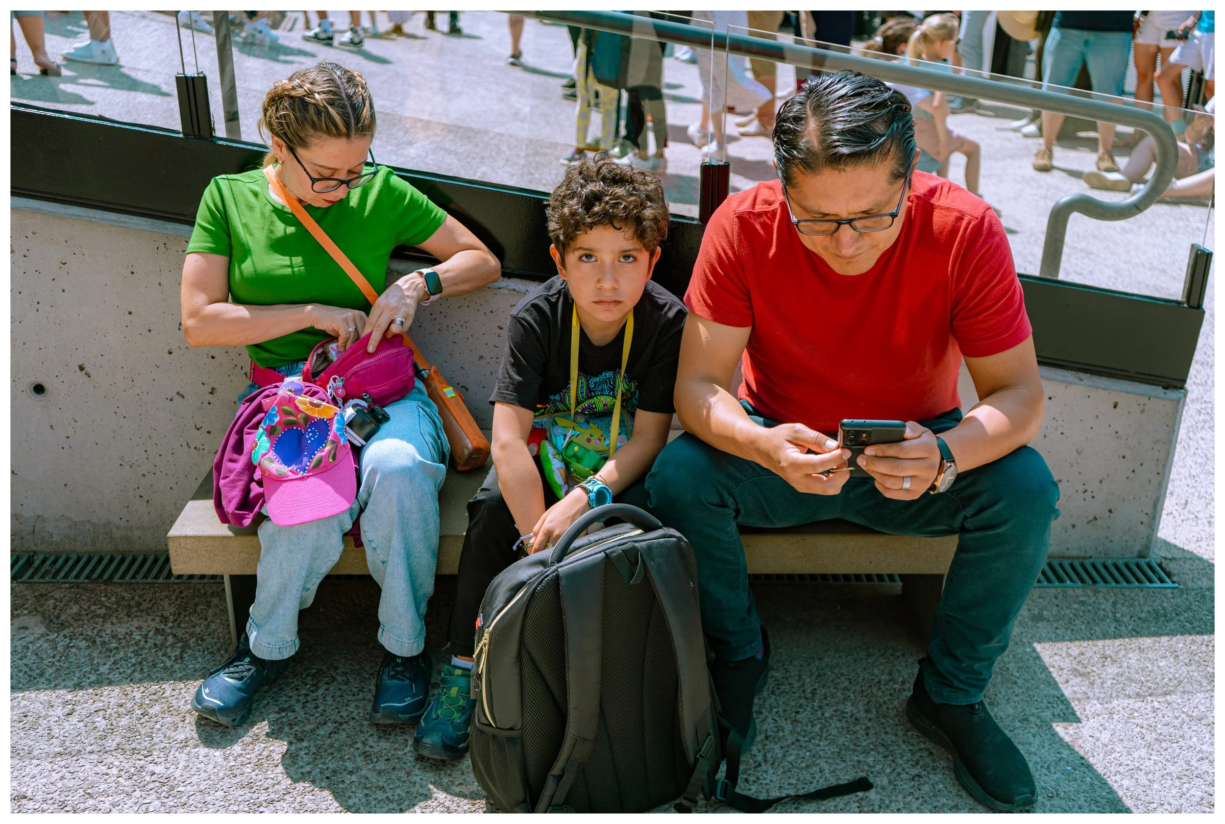 A family of three sitting on a bench at an outdoor location, possibly an airport. The woman on the left is wearing a green shirt and glasses, looking into her pink purse. The young boy in the middle has curly hair, wears a black shirt with colorful d