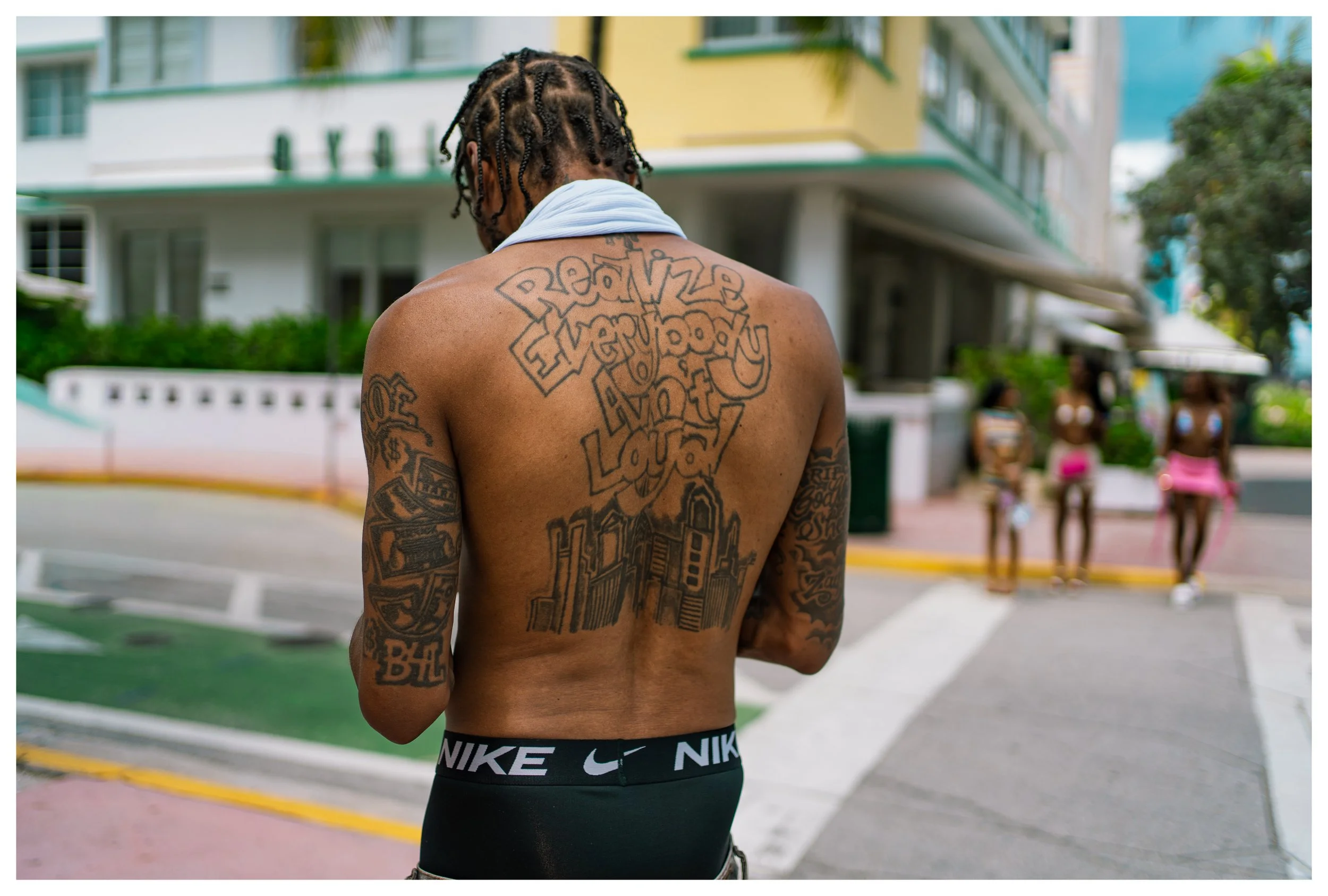 A shirtless man with tattoos on his back and arms stands outdoors with three women in swimsuits in the background. The man has braided hair, a white towel around his neck, and is wearing Nike underwear.
