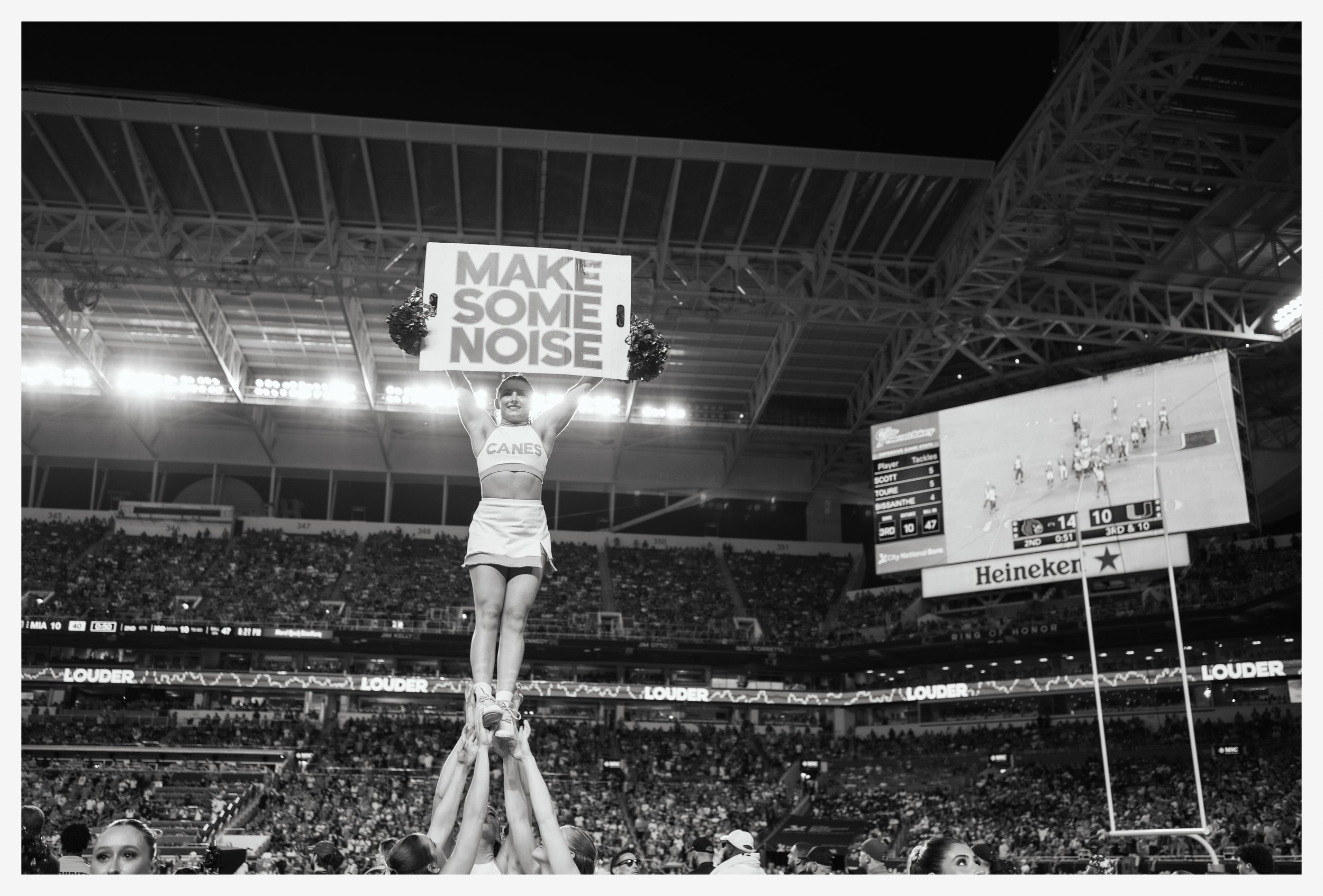 A cheerleader holding a sign that says 'MAKE SOME NOISE' during a football game in a stadium.