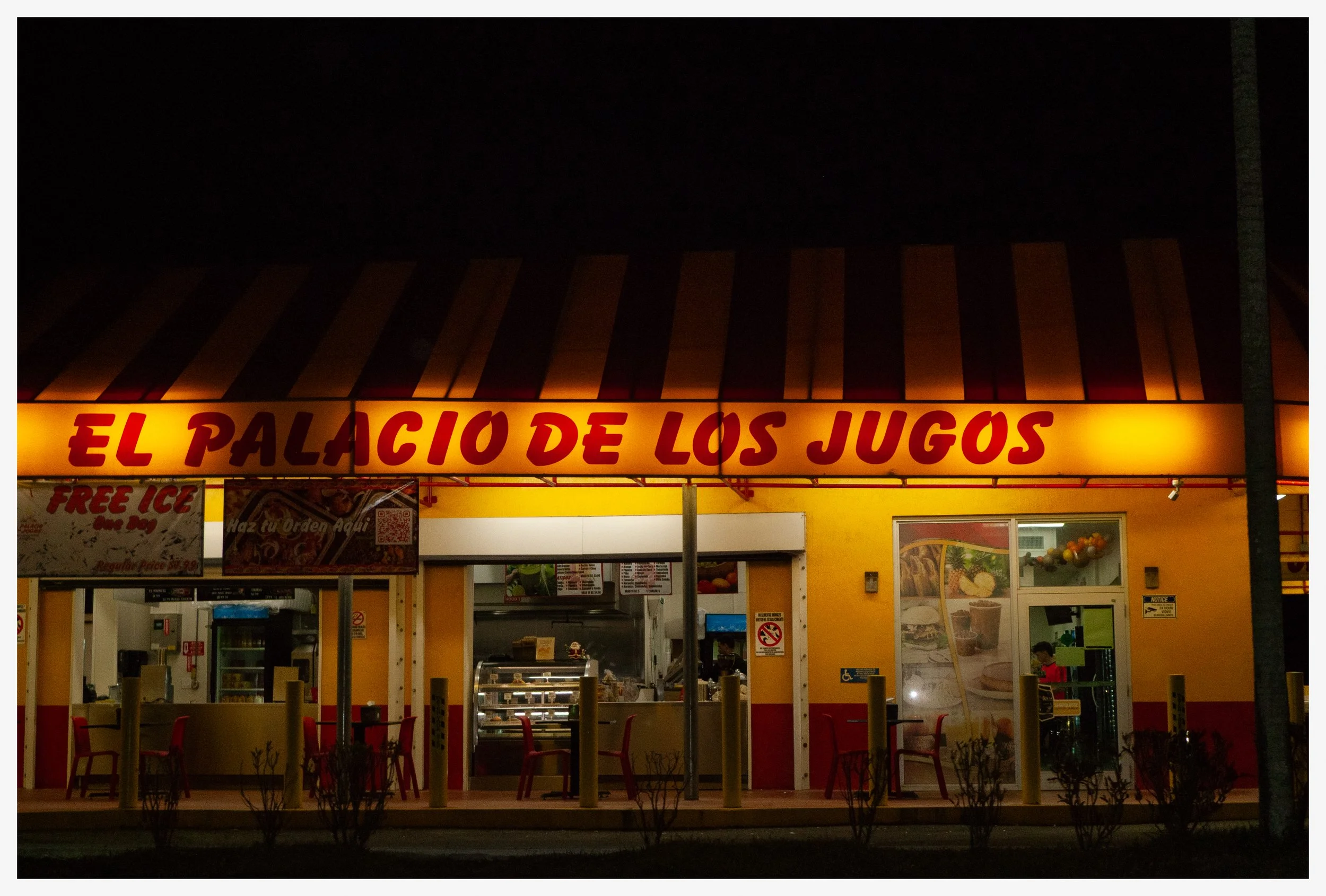 Nighttime view of a brightly lit fast-food restaurant called 'El Palacio de los Jugos' with yellow and red signage, outdoor seating, and promotional posters on the window.