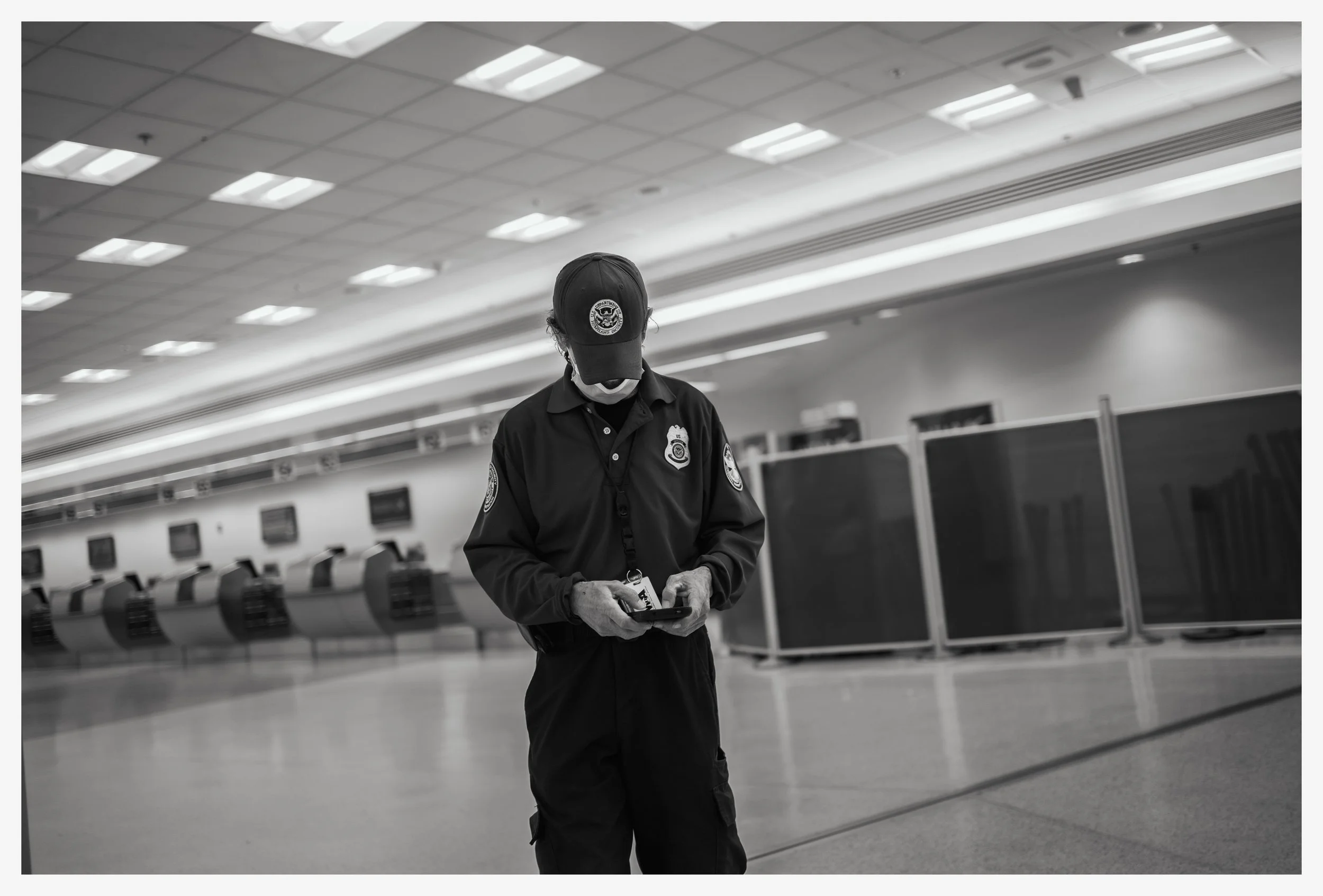 A security officer in uniform and face mask standing in an empty airport terminal using a handheld device.