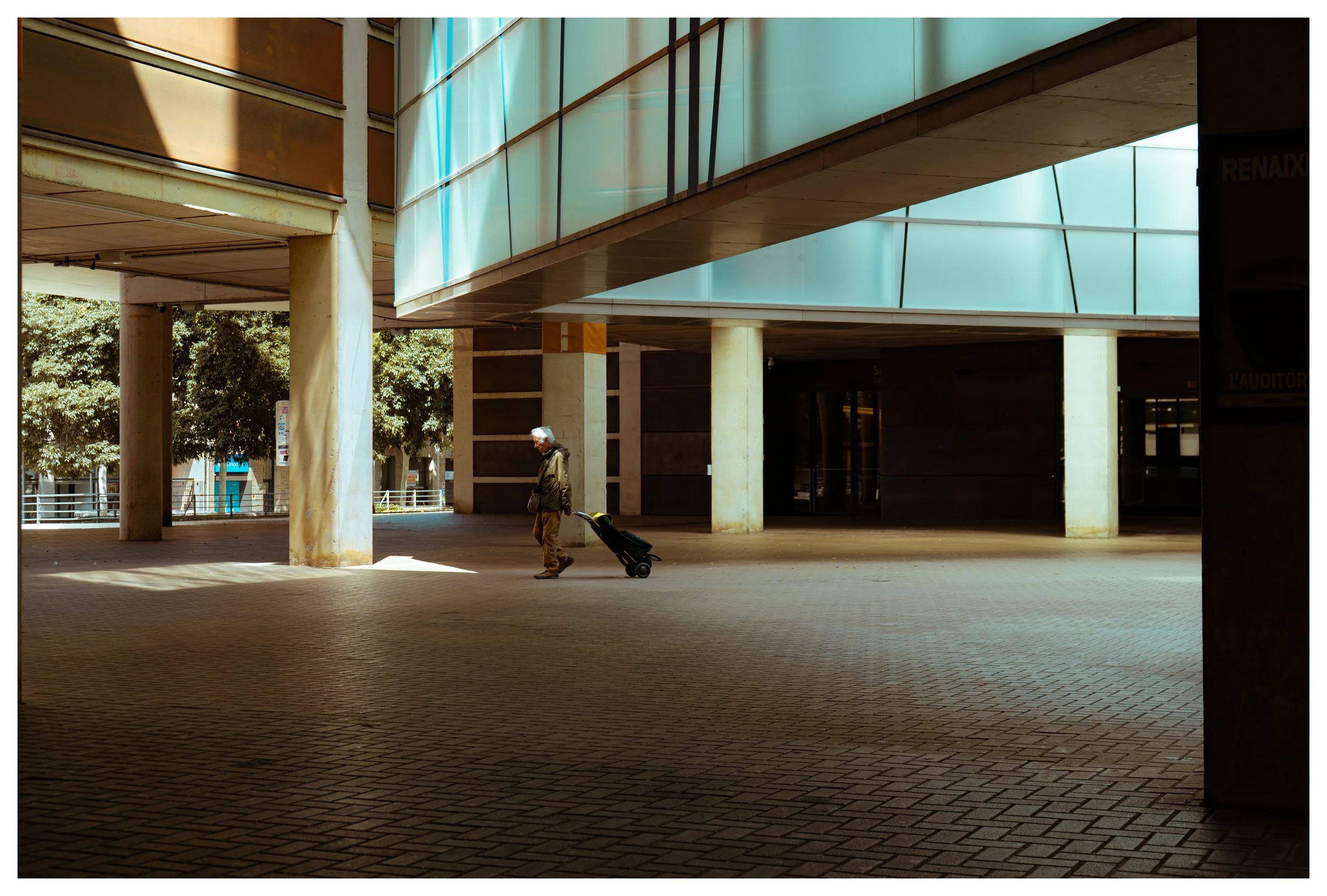 Man walking with a rolling suitcase under a modern building with large glass panels and concrete columns, shadows cast on brick-paved ground.