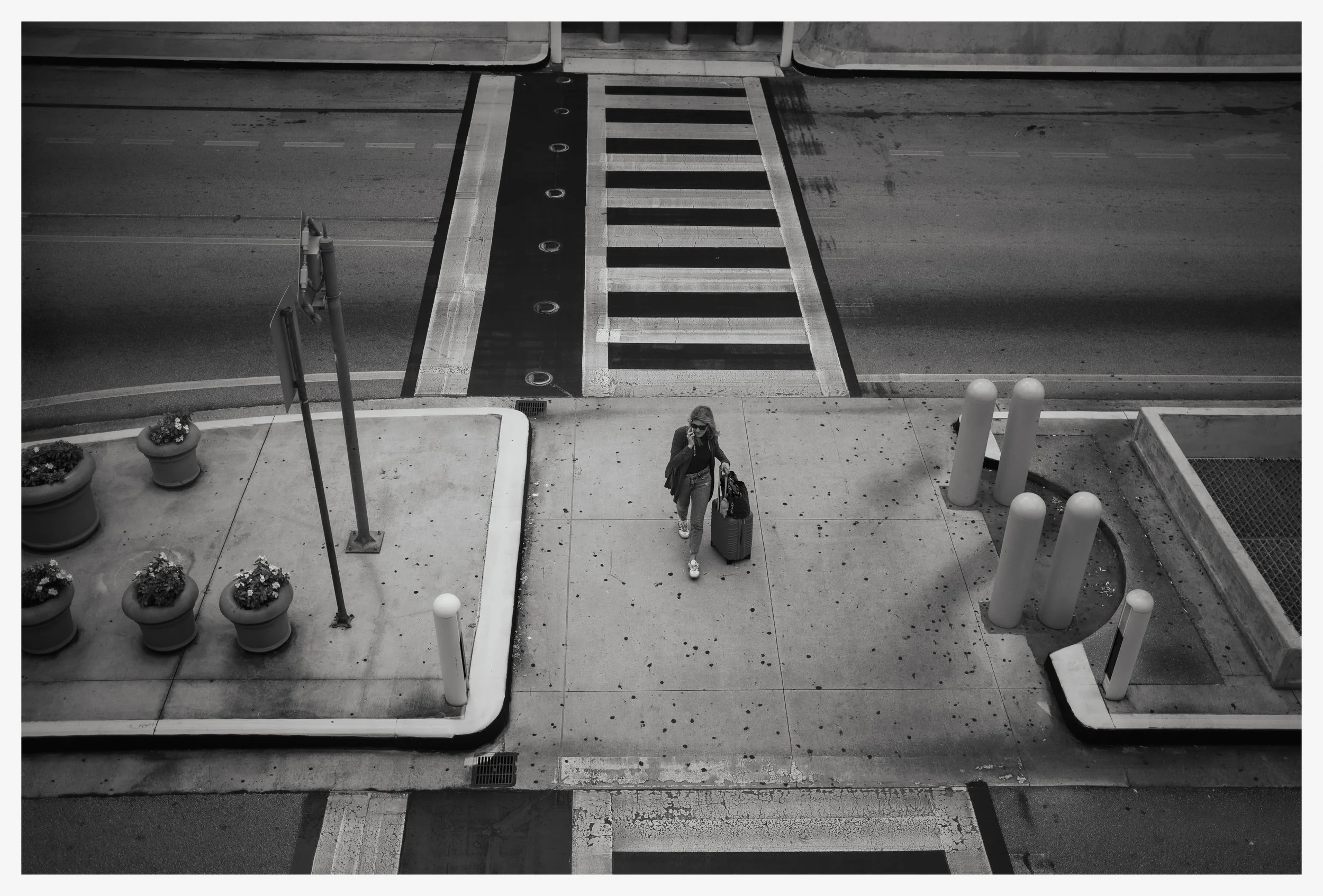 A woman with sunglasses and a backpack walks on the sidewalk, pulling a rolling suitcase, near a crosswalk on an empty street.