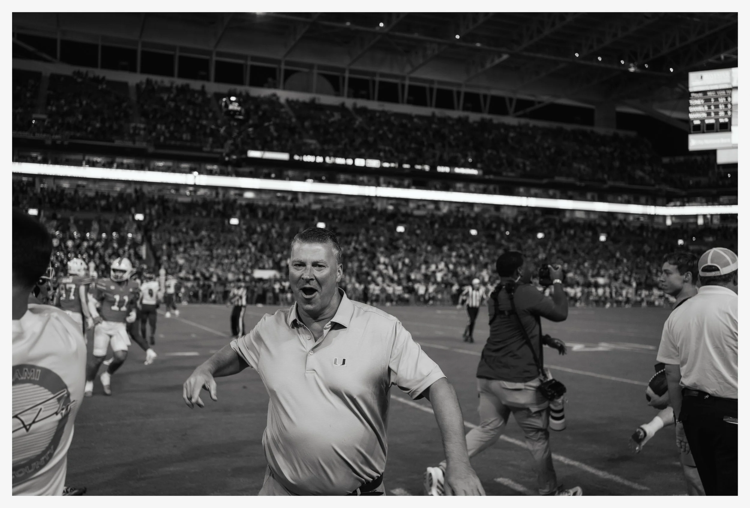 A football coach celebrating on the sideline of a football field during a game at a packed stadium, with players and photographers around him.