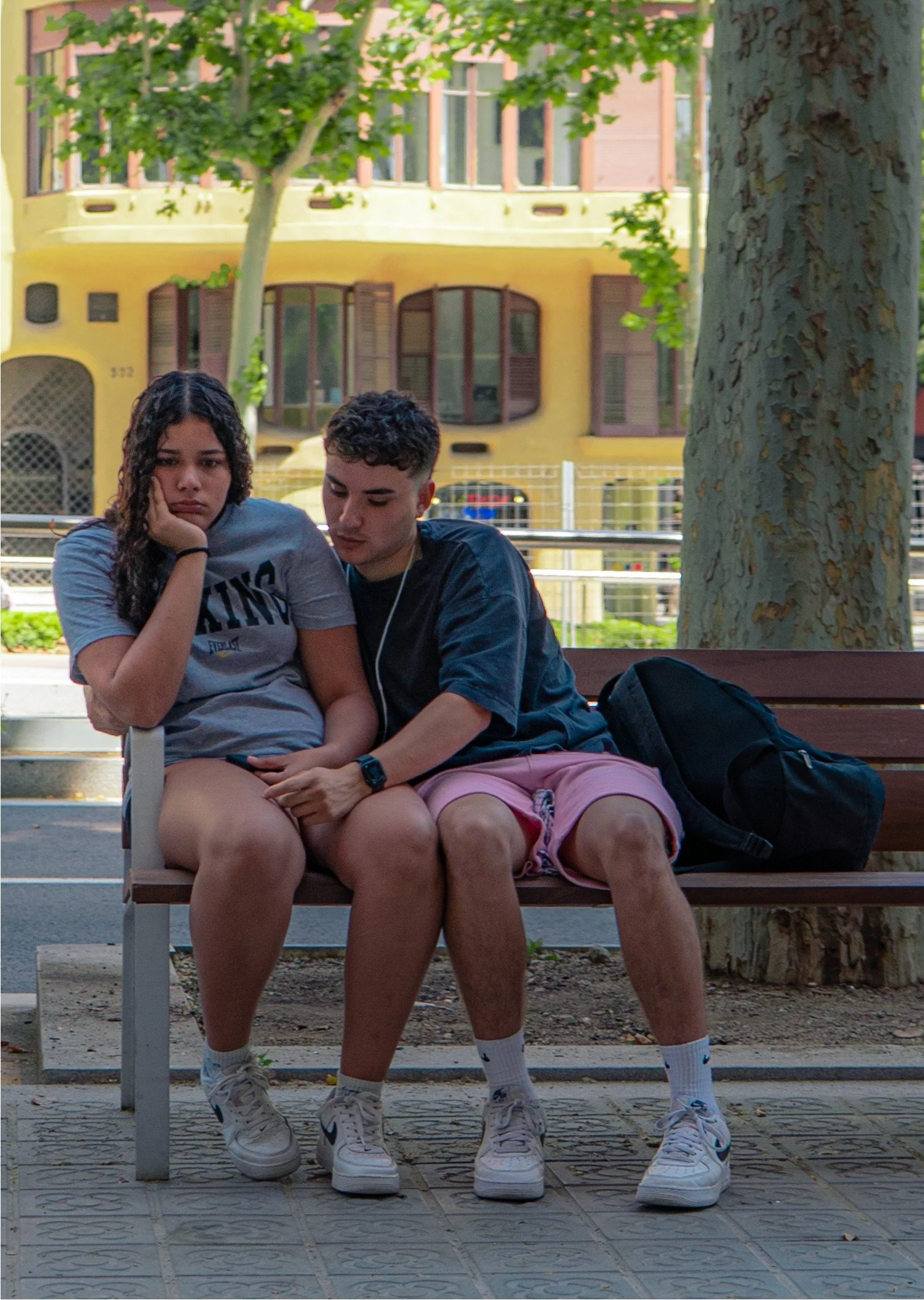 A teenage girl and boy sitting closely on a park bench, both looking down with somber expressions. The girl has curly dark hair, wearing a gray t-shirt and white sneakers, with her hand resting on her cheek. The boy has short curly hair, wearing a black shirt and pink shorts with white Nike sneakers, with his arm around the girl. A black backpack is next to them on the bench, and there are trees and a yellow apartment building in the background.