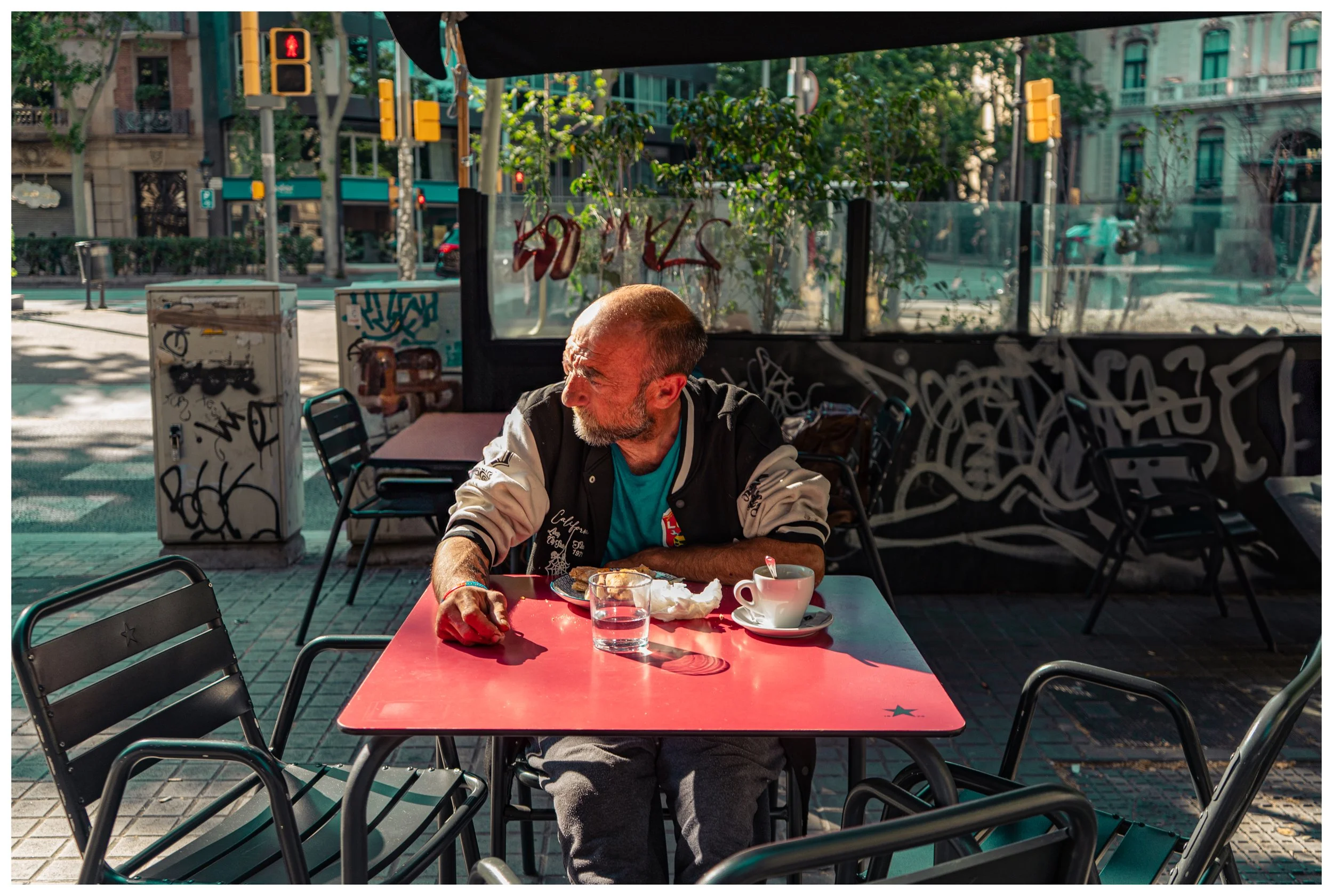 An older man with a beard and short hair sitting at a table outside a cafe, wearing a black and white jacket over a turquoise shirt, looking to his left. The table has a glass of water, a coffee cup, and some food. The sun is casting shadows, and the