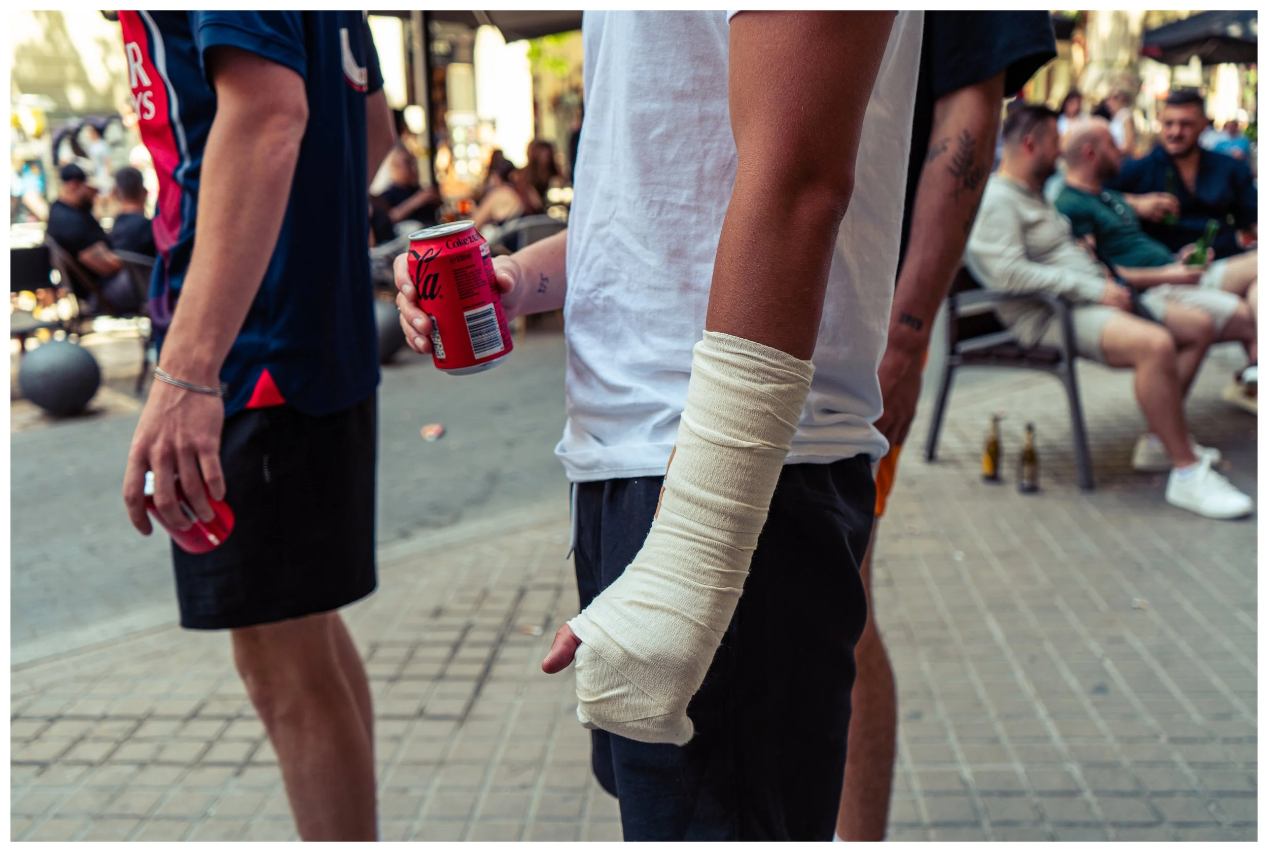People standing on a busy street, some holding soda cans, with others sitting on chairs and socializing in the background.
