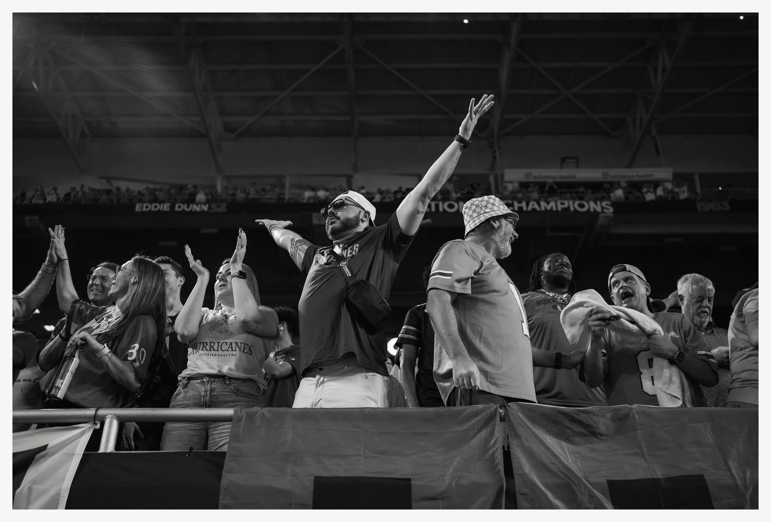 Group of diverse sports fans on stadium stands celebrating, cheering, and reaching out, with some holding towels or drinks, under a large indoor stadium roof, in black and white.