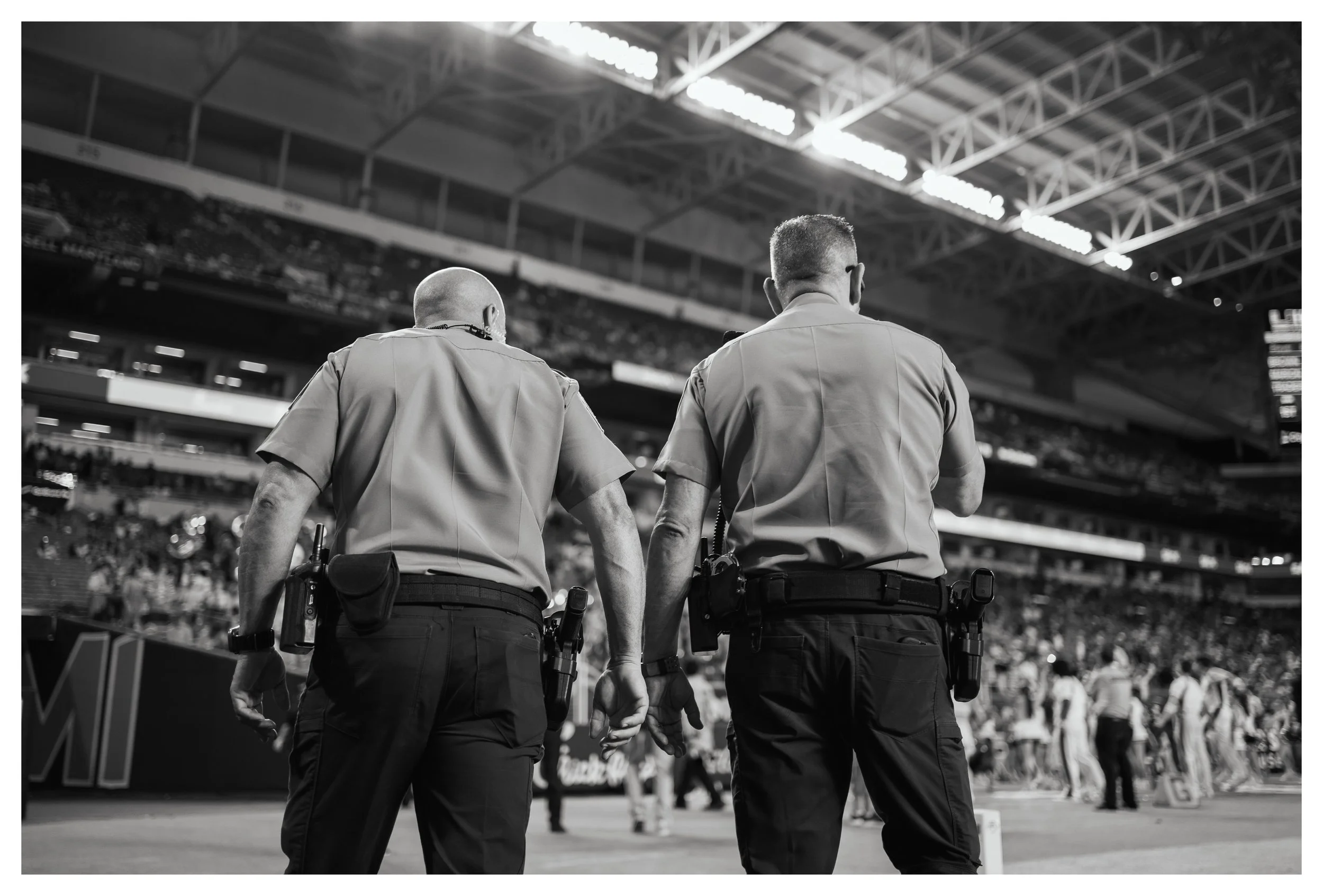 Two police officers holding hands at a sports event in a stadium, seen from behind.