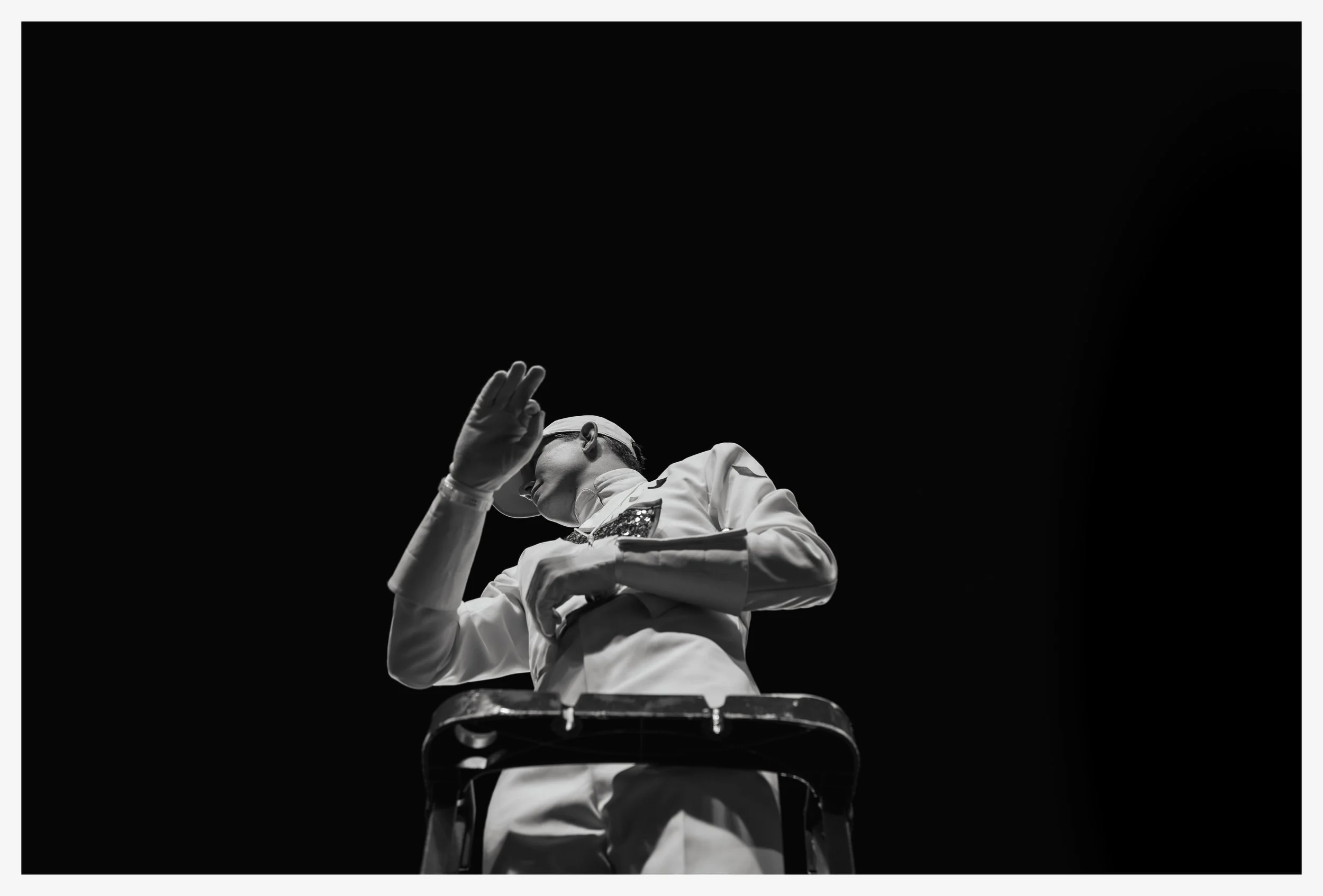 Black and white photo of a person in white attire, wearing glasses and gloves, standing on a chair against a dark background, with one hand raised to their forehead.