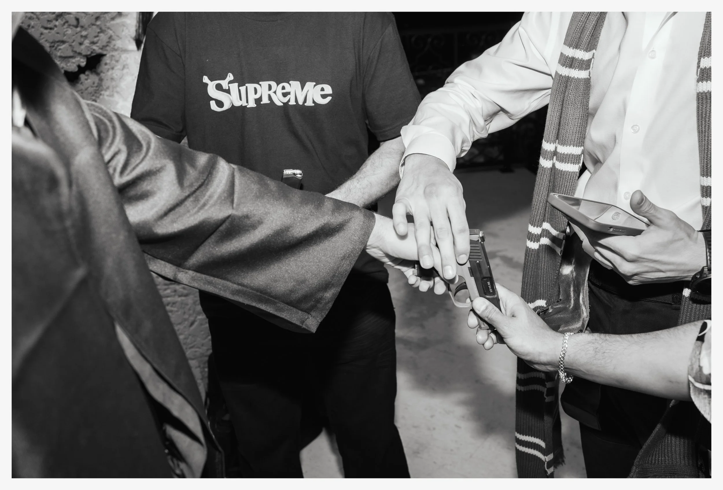 A group of people engaged in a gun safety demonstration, with one person pointing a revolver into the palm of another person's hand while others observe and hold a smartphone, in a black and white photo.