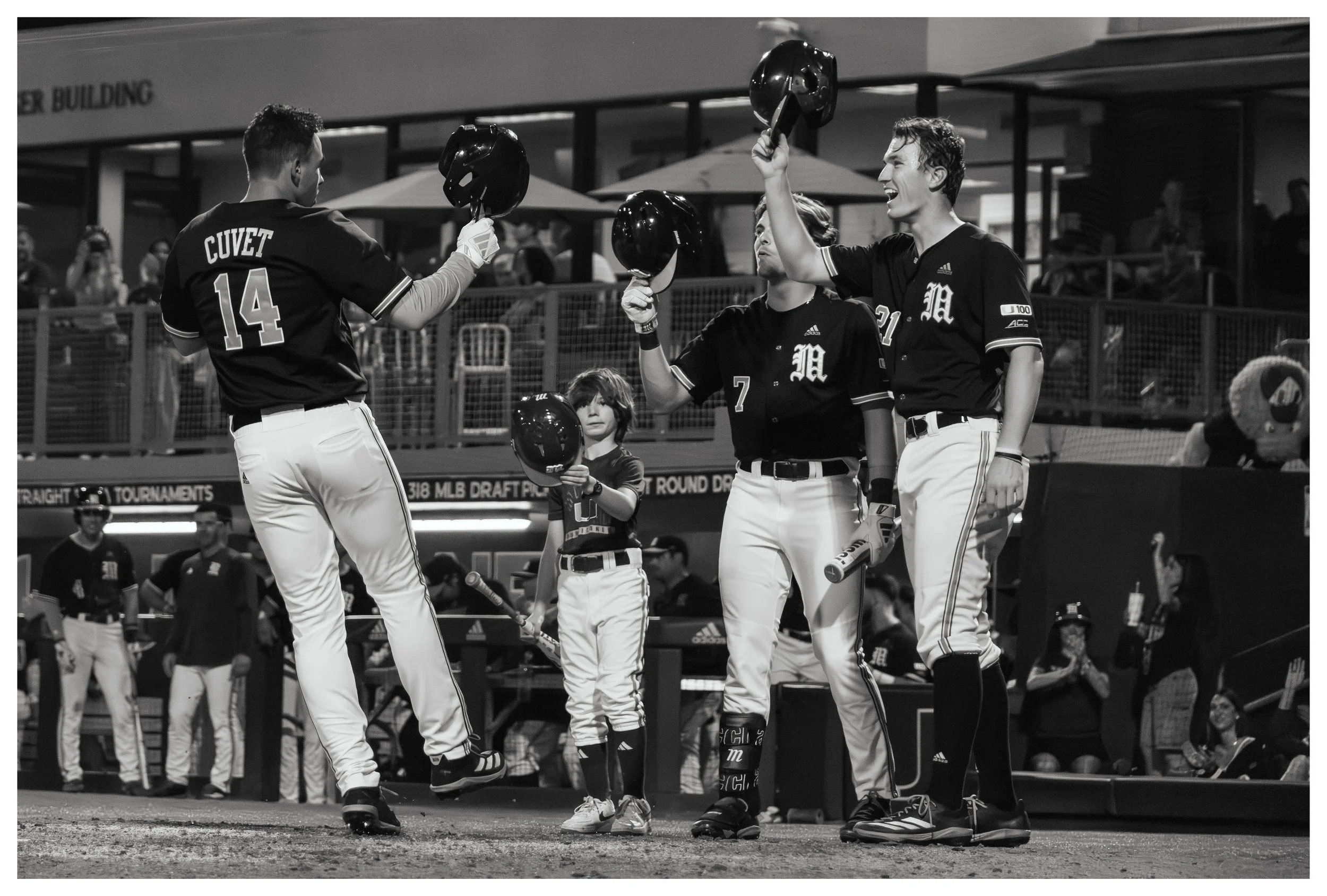 Baseball players celebrating on the field, some holding their helmets in the air. A young player stands between two, holding his helmet with a serious expression. Other players and spectators are visible in the background.