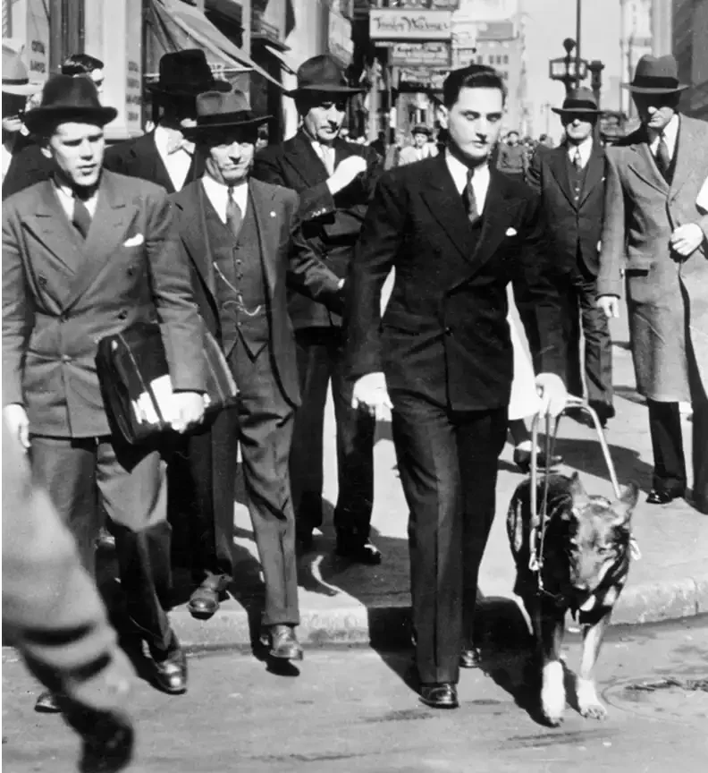 Black-and-white photo of Morris Frank holding Buddy's harness as they walk a busy New York street, surrounded by suited men watching closely.