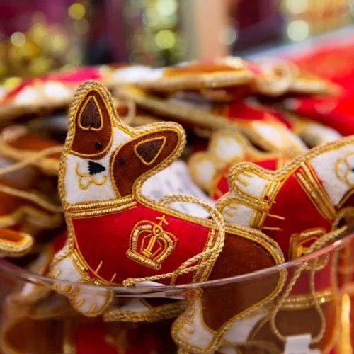 Felt Corgi-shaped Christmas ornaments dressed in red royal attire with gold trim and crown emblems, displayed in a bowl, celebrating the breed's royal connection.