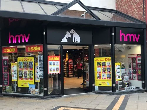 Modern HMV store in Coventry with bright sale posters, pink logo signage, and a simplified white silhouette of Nipper the dog listening to a gramophone above the entrance.