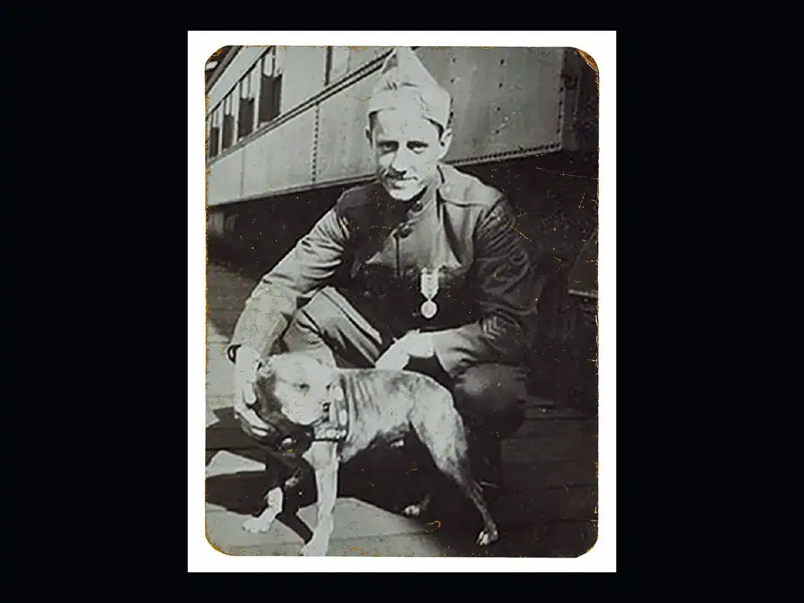 A World War I soldier crouches beside a Boston terrier mix dog near a train car, the dog is wearing a collar, both posing calmly for the camera.