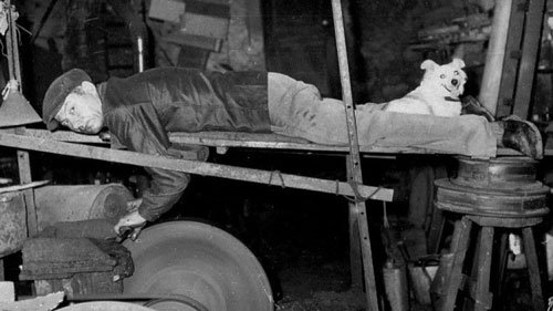 Vintage photo of a French knife grinder lying on a wooden plank while sharpening a blade, with a small dog lying on his legs for warmth.