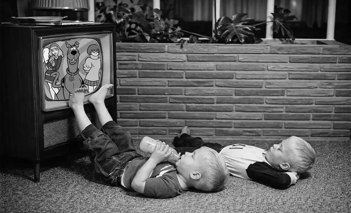 Black-and-white photo of two young boys lying on a carpet watching a vintage TV showing Scooby-Doo; one drinks from a bottle with feet resting against the set.