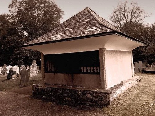 A stone mausoleum with a pyramidal slate roof and iron-barred walls stands in a churchyard in Buckfastleigh, Devon