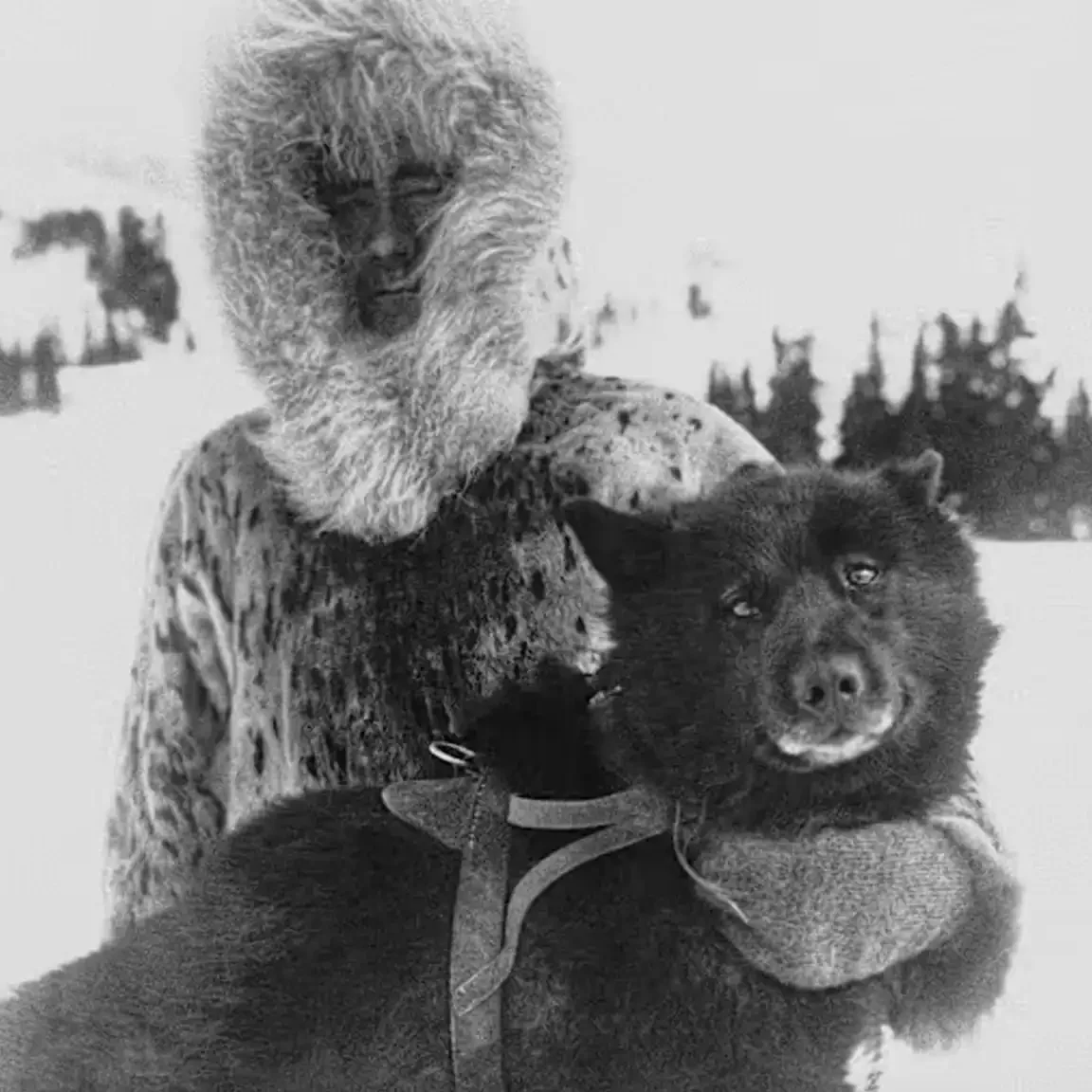 A black-and-white close-up photo of a person in a thick fur-hooded parka holding a dark, fluffy dog's head and harness close to their chest in the snow