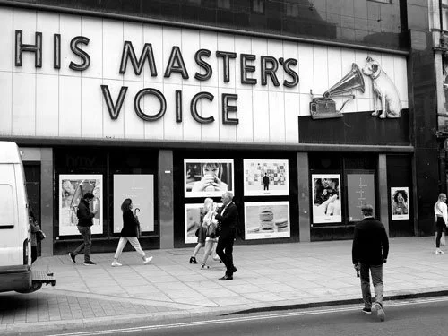 Black and white photo of the HMV store on Oxford Street, London, featuring the large His Master’s Voice sign with Nipper and a gramophone above the shopfront as people walk past.