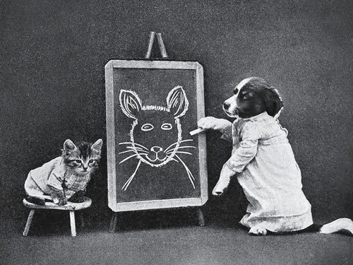 A dressed puppy points to a chalk drawing of a mouse on a blackboard while a kitten, also dressed, sits on a stool watching in this staged scene from The Little Folks of Animal Land.