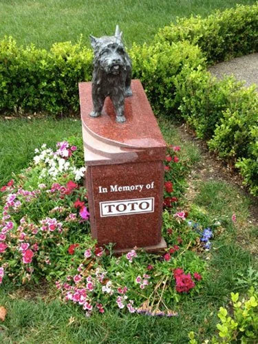 Bronze statue of Toto stands atop a red granite memorial reading "In Memory of TOTO," surrounded by flowers at Hollywood Forever Cemetery.