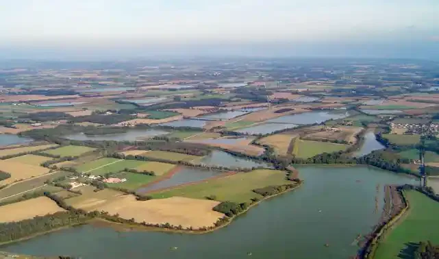 An aerial view of flat farmland cut through by dozens of shallow ponds and lakes, with hedgerows, plowed fields and scattered farm buildings stretching to a hazy horizon.