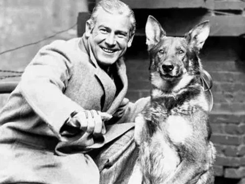 Black-and-white photo of a smiling man in a coat crouching beside a German shepherd, both facing the camera.