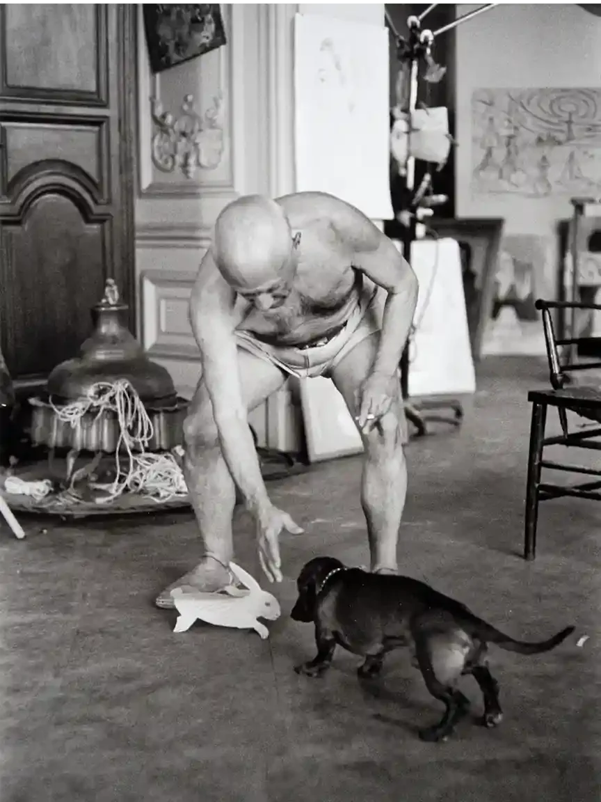 A shirtless elderly man stoops low to place a white cardboard rabbit on the floor. A small black dachshund approaches it cautiously, tail raised, in a cluttered studio.