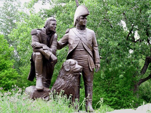Bronze monument of Lewis and Clark standing with their Newfoundland dog Seaman in a wooded park setting.