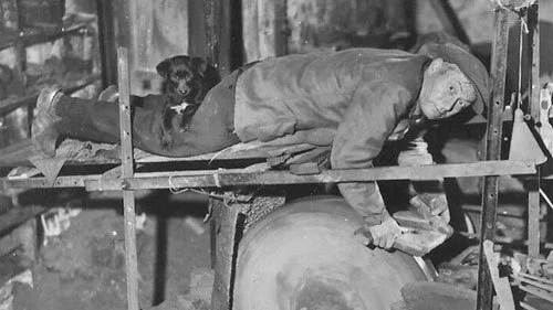 Black and white photo of a French knife grinder lying over a sharpening wheel, with a small dog resting on his legs for warmth in a cluttered workshop.