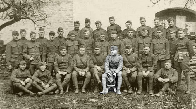 Sepia group photo of uniformed WWI soldiers posed in rows outdoors, with a man in the center holding a small dog. Public domain, via Wikipedia Commons.