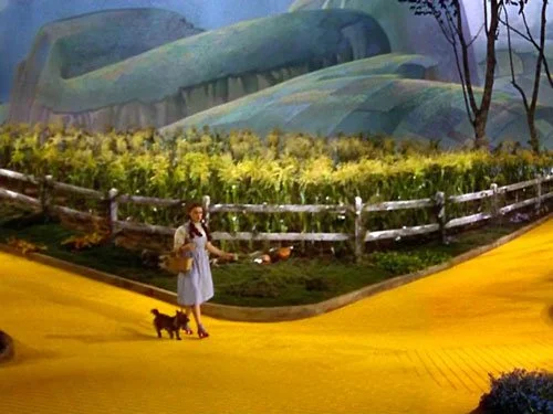 Dorothy and Toto stand at a fork in the Yellow Brick Road, surrounded by cornfields and painted hills, in a vibrant scene from The Wizard of Oz (1939).