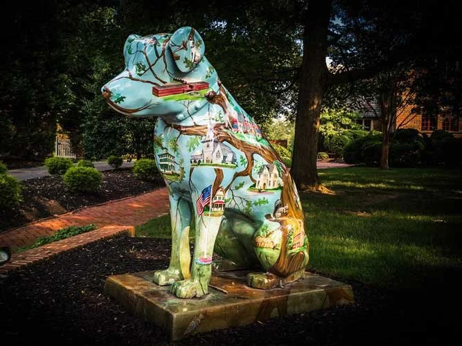 Colourful outdoor statue of Nipper the dog, painted with scenes of American landmarks and landscapes, displayed on a brick path in a landscaped garden.