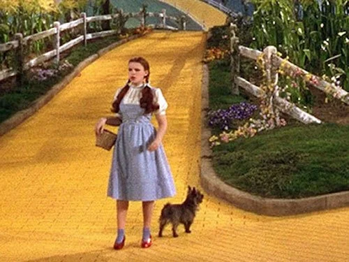 Dorothy stands on the Yellow Brick Road with Toto at her side, holding a basket and looking ahead in a colourful scene from The Wizard of Oz (1939).