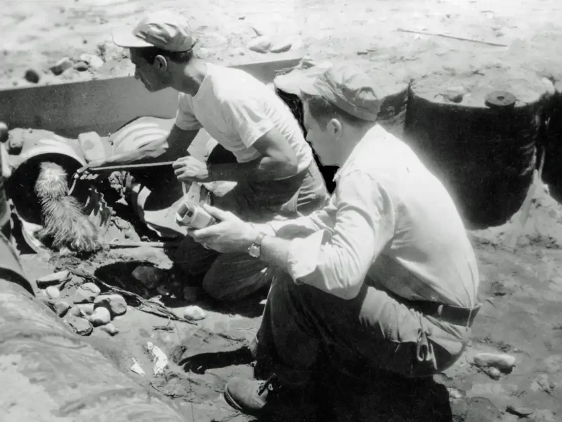 Black and white photo: two soldiers crouch over a pipe opening in sandy ground, guiding a small terrier toward the entrance.