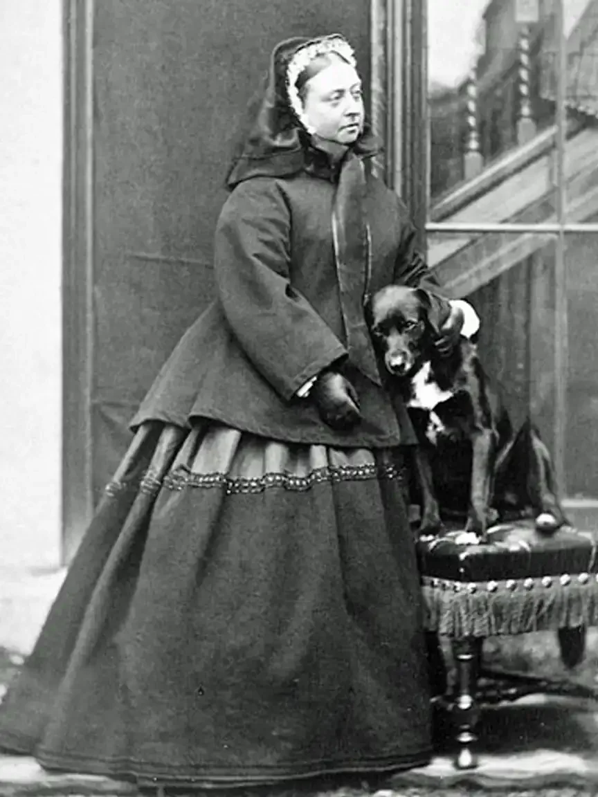 Queen Victoria stands beside her dog perched on a small upholstered table, both posed formally in a Victorian studio setting, wearing period clothing and conveying quiet intimacy.