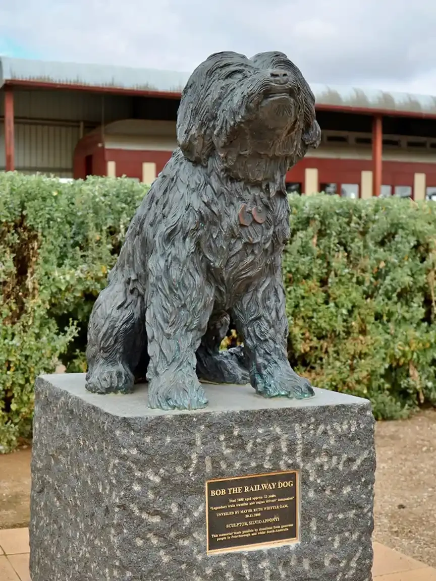 A bronze statue of a shaggy seated dog wearing a collar tag stands on a rough granite plinth outside a red-and-cream railway building. A plaque at the base reads Bob the Railway Dog.