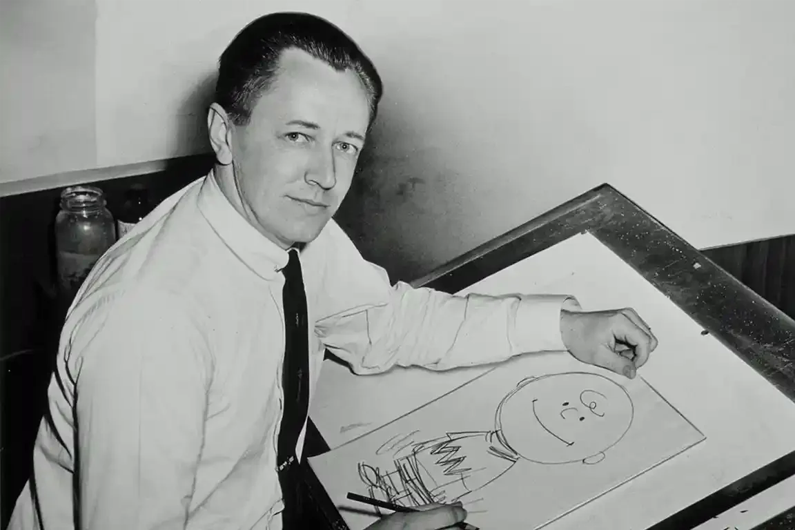 Black-and-white photo of a man in a white shirt and dark tie seated at a tilted drawing board, turned toward the camera. On the paper is a pencil sketch of a round-headed smiling boy.
