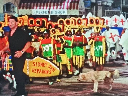 A festive street parade with colorful costumes wearing 007 hats; a men in black on the left hand side of a dog peeing on the right of the street. Scene from James Bond movie Thunderball 1965