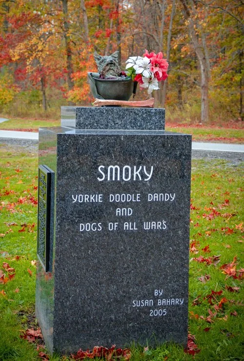 Blue granite memorial stone topped with a bronze sculpture of Smoky in a GI helmet, inscribed "Smoky, Yorkie Doodle Dandy and Dogs of All Wars, by Susan Bahary, 2005."