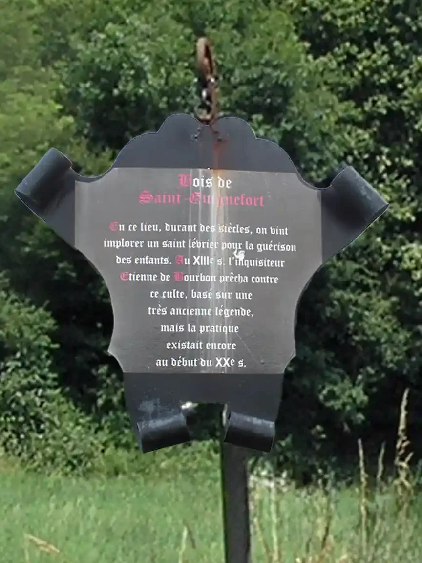 A dark metal sign shaped like a shield hangs from a post in a green field backed by trees. Red and white French lettering is headed Bois de Saint-Guinefort.