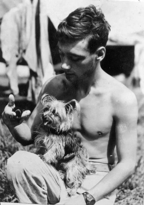 Black and white photo: young shirtless soldier sits outdoors cradling a small Yorkshire Terrier in his lap, looking down at her.