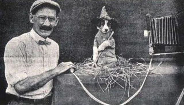 Photographer Harry Whittier Frees poses beside a costumed puppy on a studio set, ready for a photo with a vintage camera in this behind-the-scenes image.