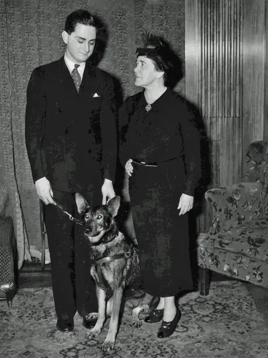 Morris Frank and Dorothy Eustis stand indoors with Buddy the German Shepherd between them; Eustis looks toward Frank, who looks down at the dog.