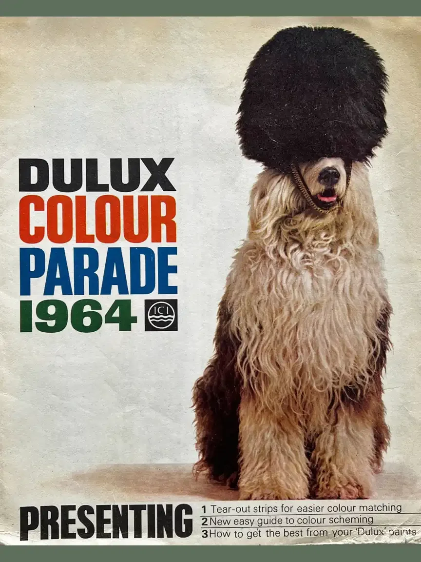 1964 Dulux Colour Parade poster showing Old English Sheepdog wearing a tall bearskin-style hat beside bold red and blue text.