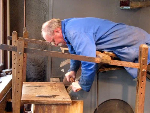 An elderly man in a blue coat lies on a wooden plank over a grinding wheel while sharpening a knife, demonstrating a traditional French knife grinder’s technique.