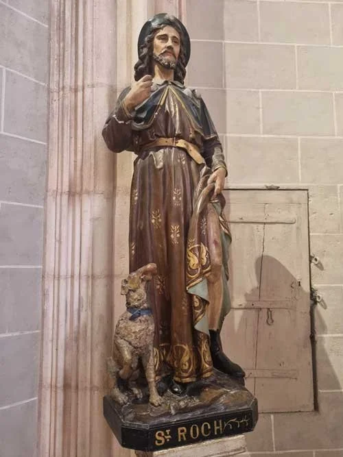 Painted wooden statue of St. Roch in pilgrim robes, one hand raised, a small dog at his feet, labeled St Roch on the base, set against a stone pillar.